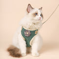 Weiss-braune Katze mit blauem Blick trägt ein grünes Katzengeschirr mit Blütenapplikation und cremefarbener Einfassung.