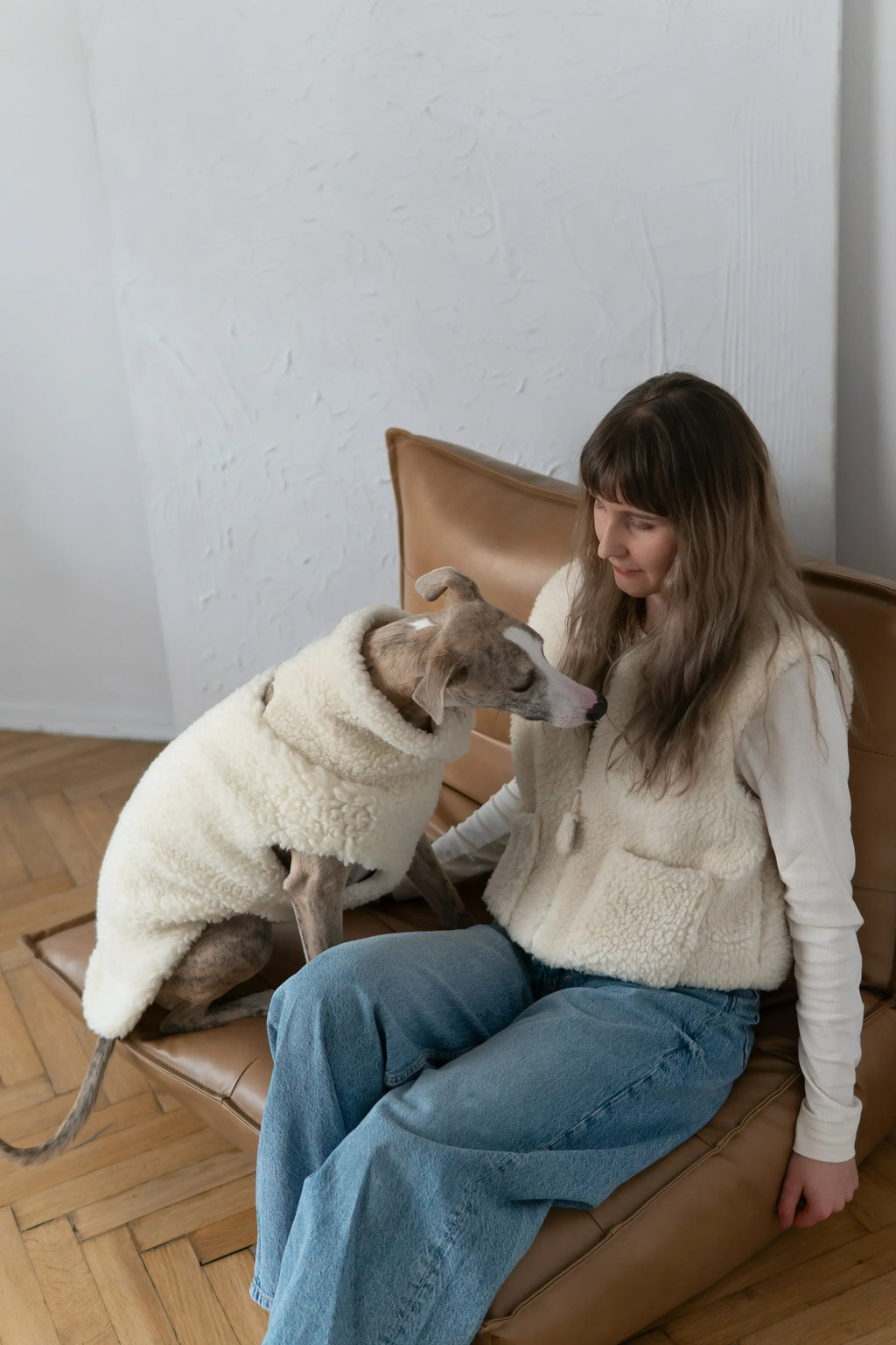 Hund in weisser Mellow-Wollweste sitzt neben einer Frau mit passender Teddyweste auf einem Ledersessel.