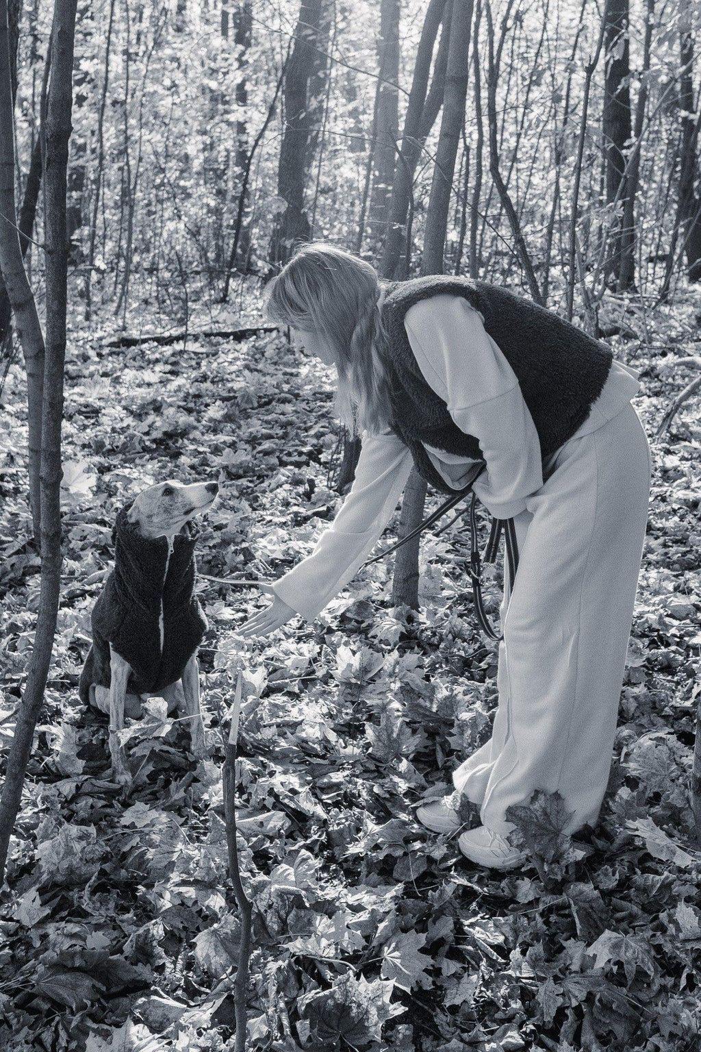 Schwarz-Weiss-Aufnahme einer Frau mit einem sitzenden Hund in einer Mellow-Wollweste im Wald.