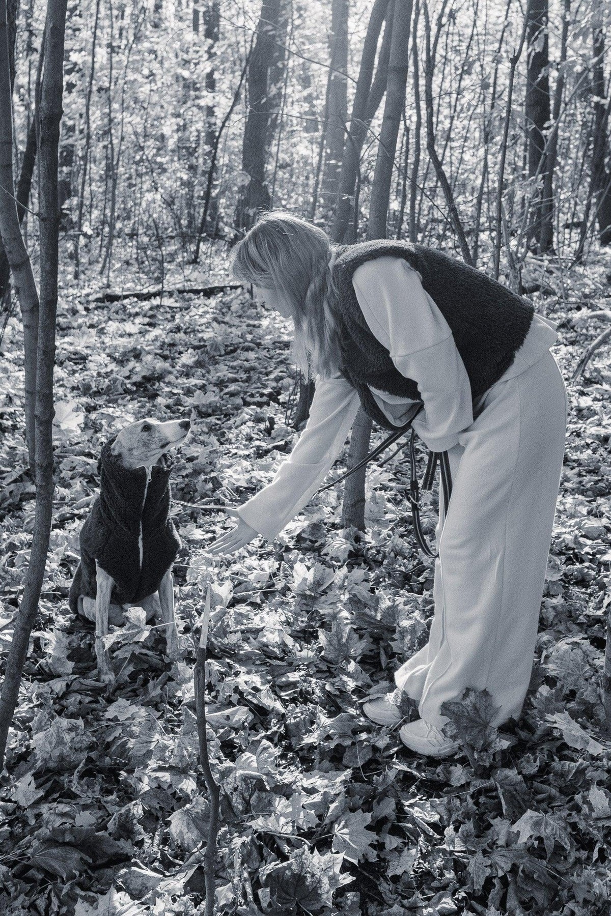 Schwarz-Weiss-Aufnahme einer Frau mit einem sitzenden Hund in einer Mellow-Wollweste im Wald.