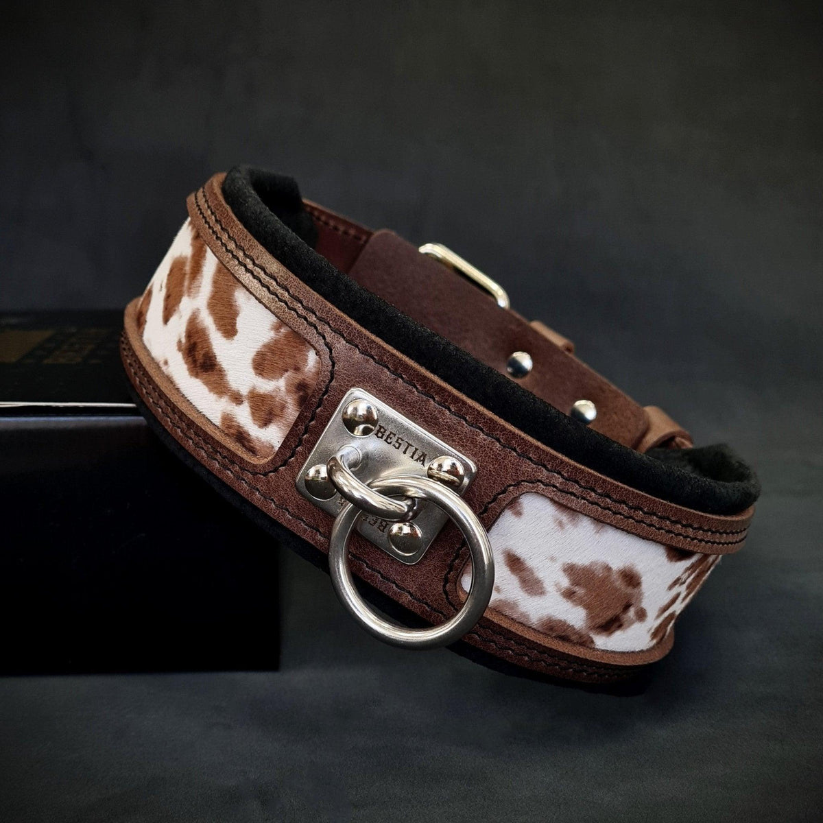 Dunkelbraunes Buffalo-Lederhalsband mit braun-weiss gefleckten Einsätzen, schwarzer Polsterung und silberfarbenem Leinenring auf dunklem Hintergrund