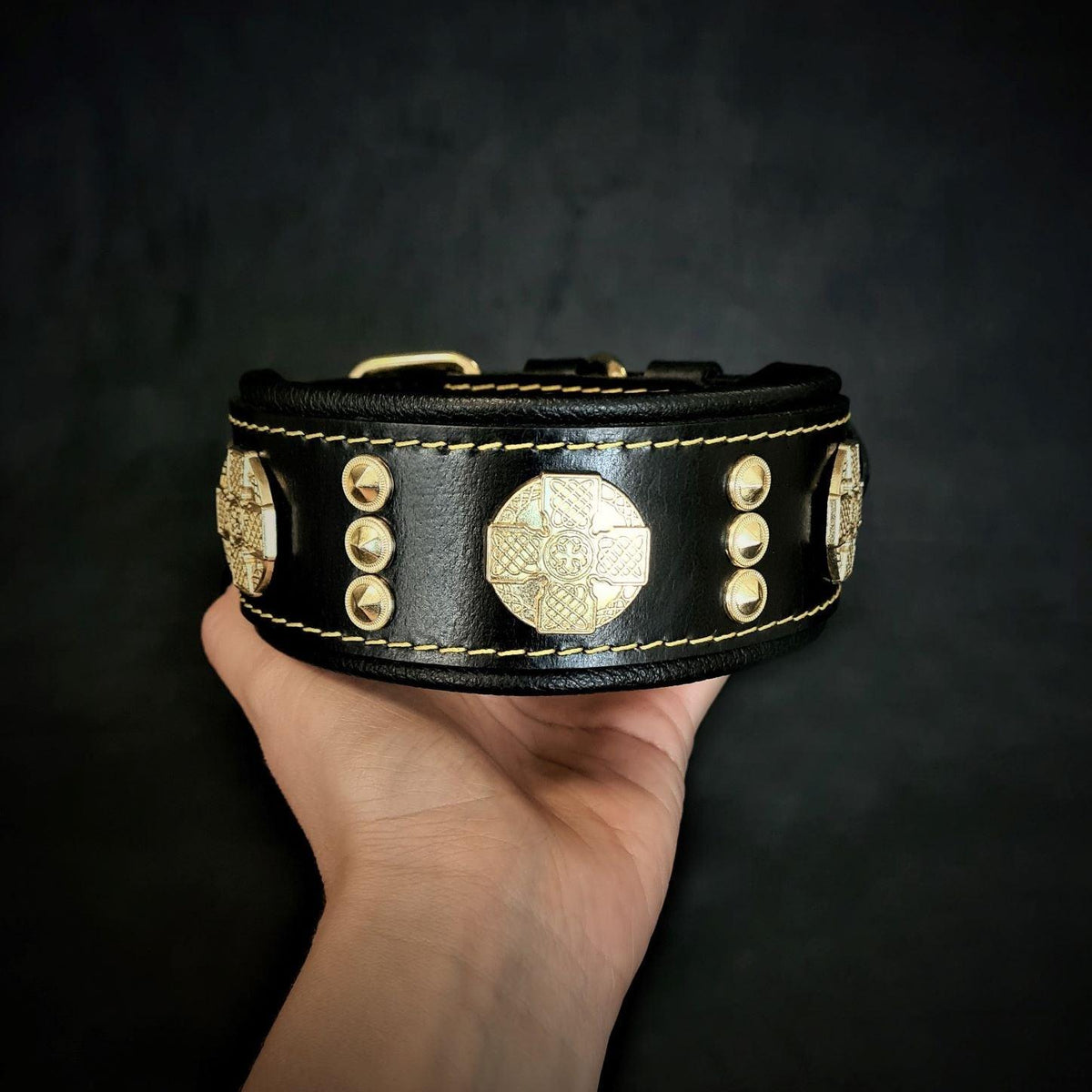 Schwarzes Maximus-Lederhalsband mit goldfarbenen Zierscheiben und Nieten sowie gelben Nähten, in einer Hand gehalten