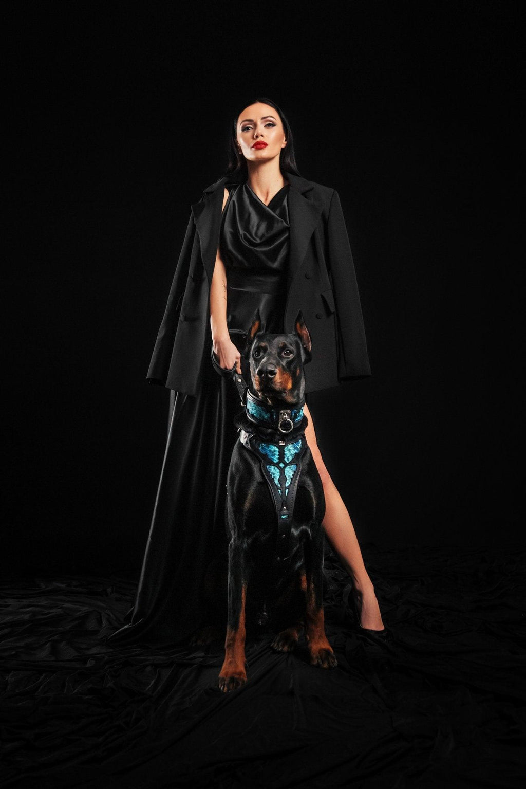 Dobermann neben einer Frau in schwarzem Kleid, mit BESTIA Morelia Python Halsband und passendem Geschirr in Schwarz/Blau.